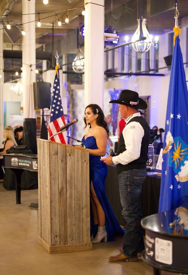 Alexia Tuttle speaking at a podium in a blue gown beside a co-host, flanked by the American and Air Force flags at the CloudDancer Gala.
